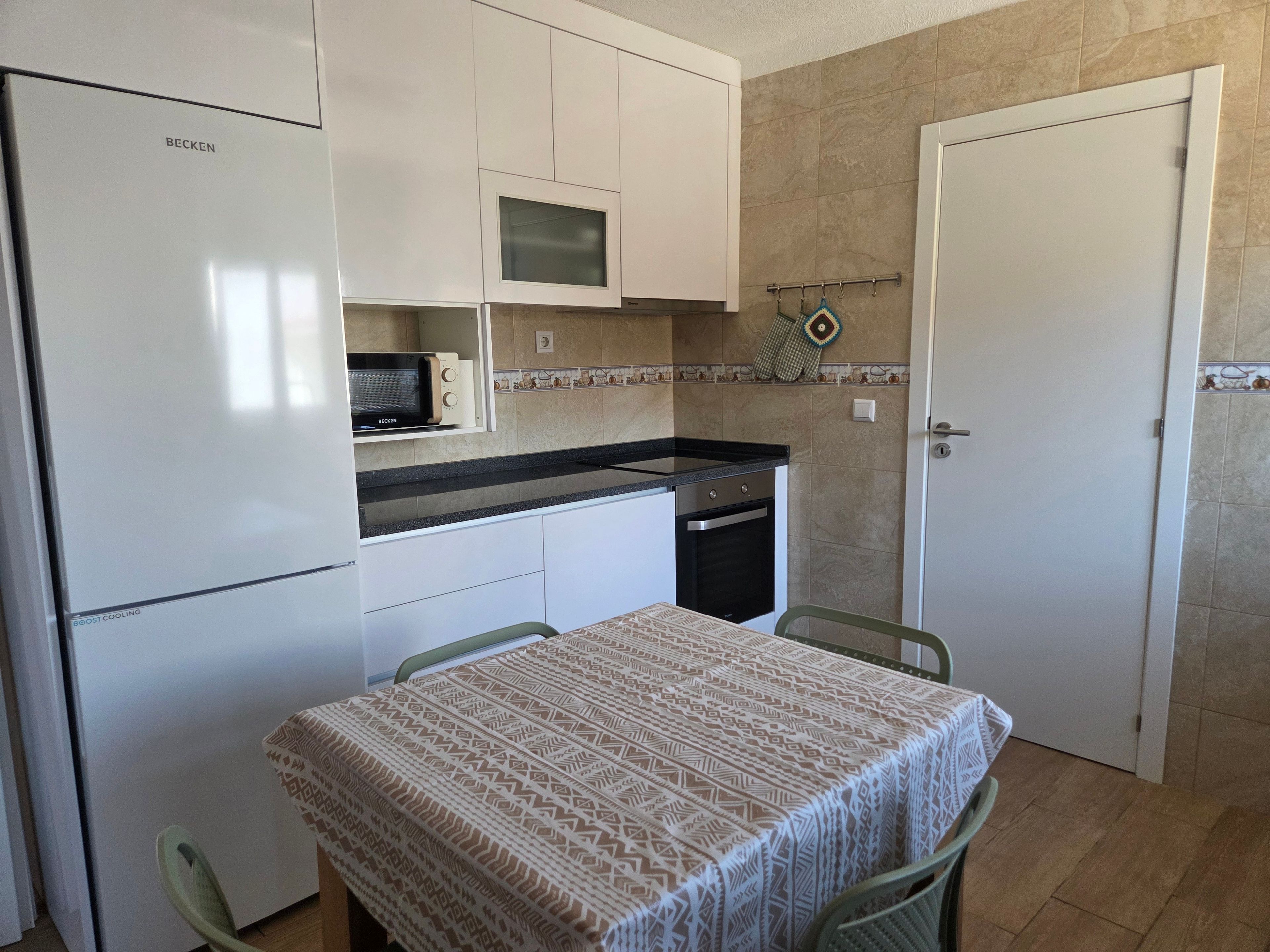 A compact kitchen with a refrigerator, microwave, stove, and dishwasher. There is a dining table with chairs and a patterned tablecloth.