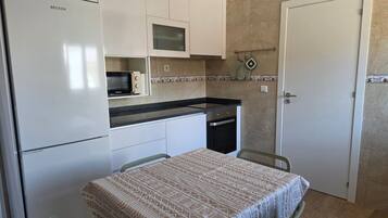A compact kitchen with a refrigerator, microwave, stove, and dishwasher. There is a dining table with chairs and a patterned tablecloth.