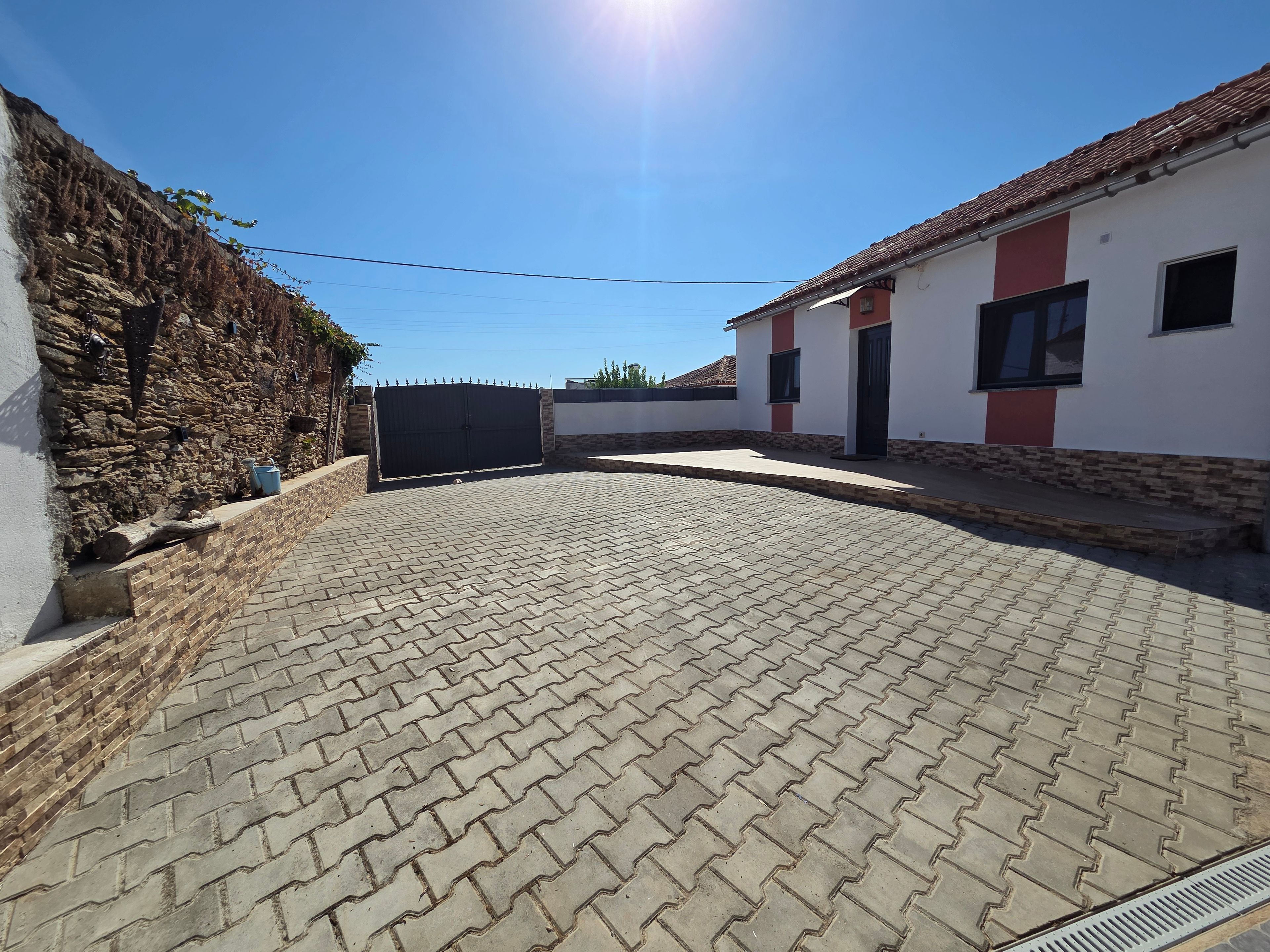 A paved courtyard with a stone wall, a metal gate, and a building with a tiled roof.
