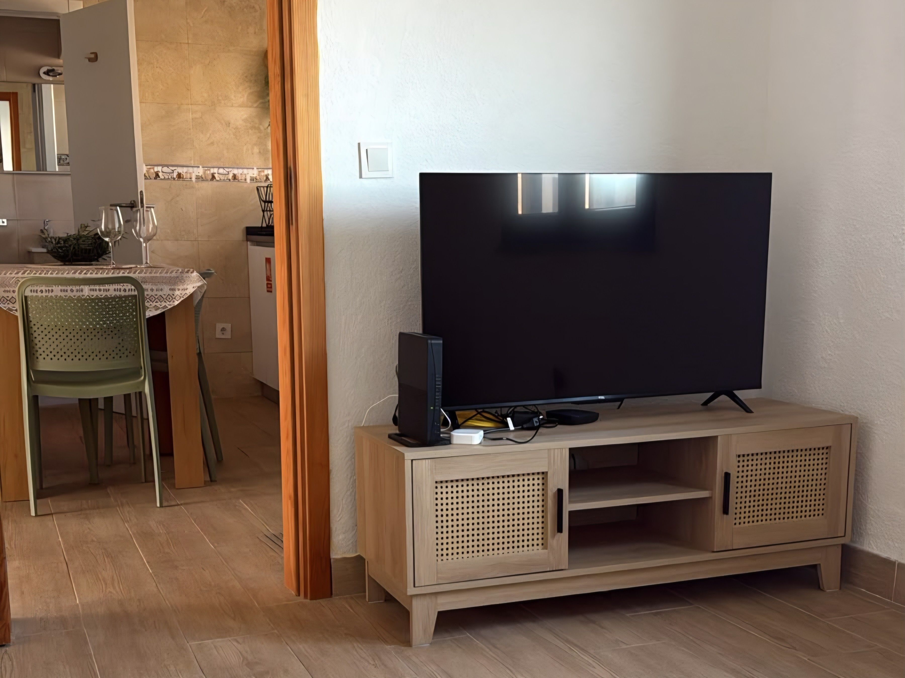 A living room with a flat-screen TV on a wooden stand, a dining area with a table and chairs, and a kitchen in the background.