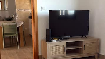 A living room with a flat-screen TV on a wooden stand, a dining area with a table and chairs, and a kitchen in the background.