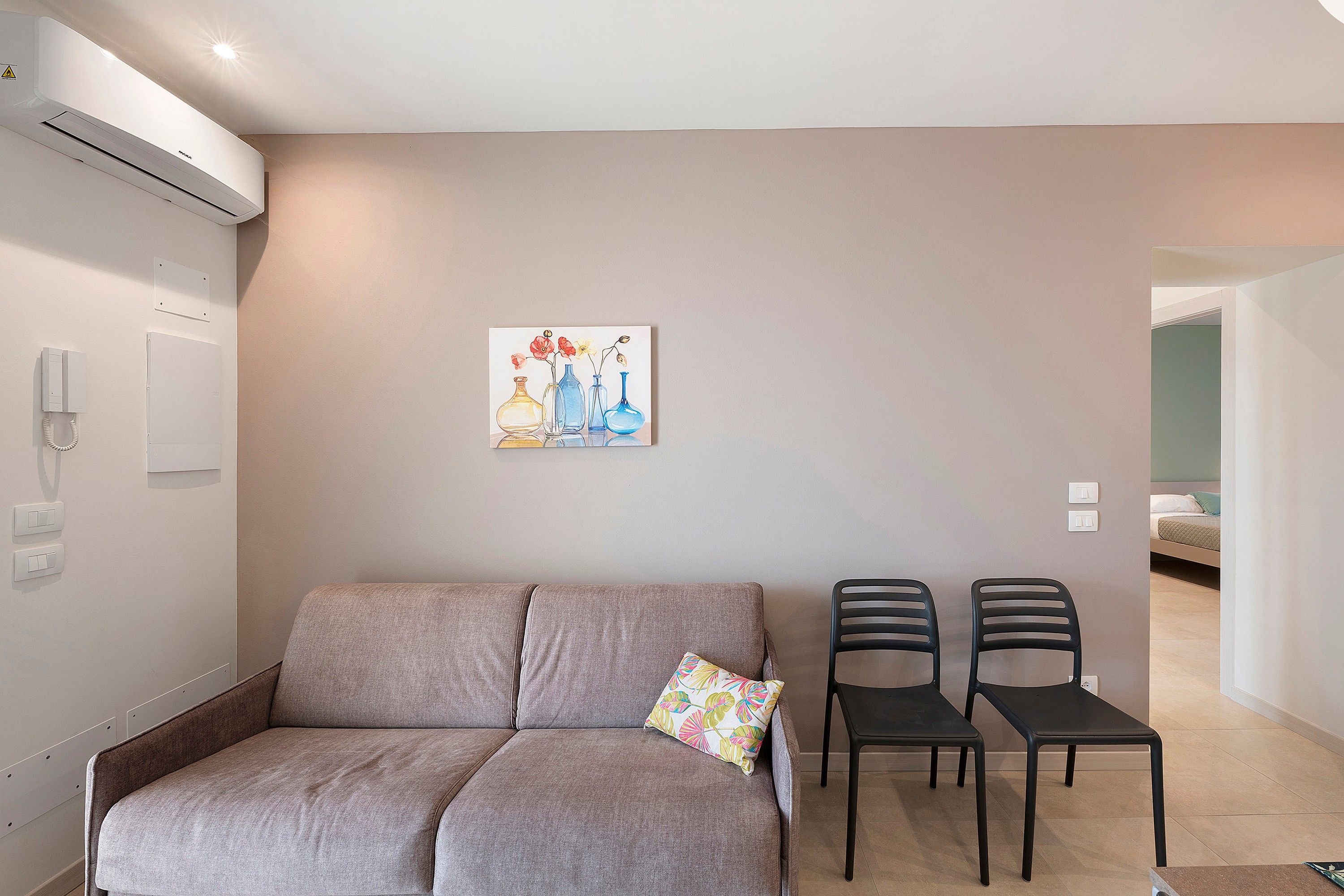 A neutral-colored room with a sofa, two chairs, a wall-mounted air conditioner, a painting, and a bed in the background.