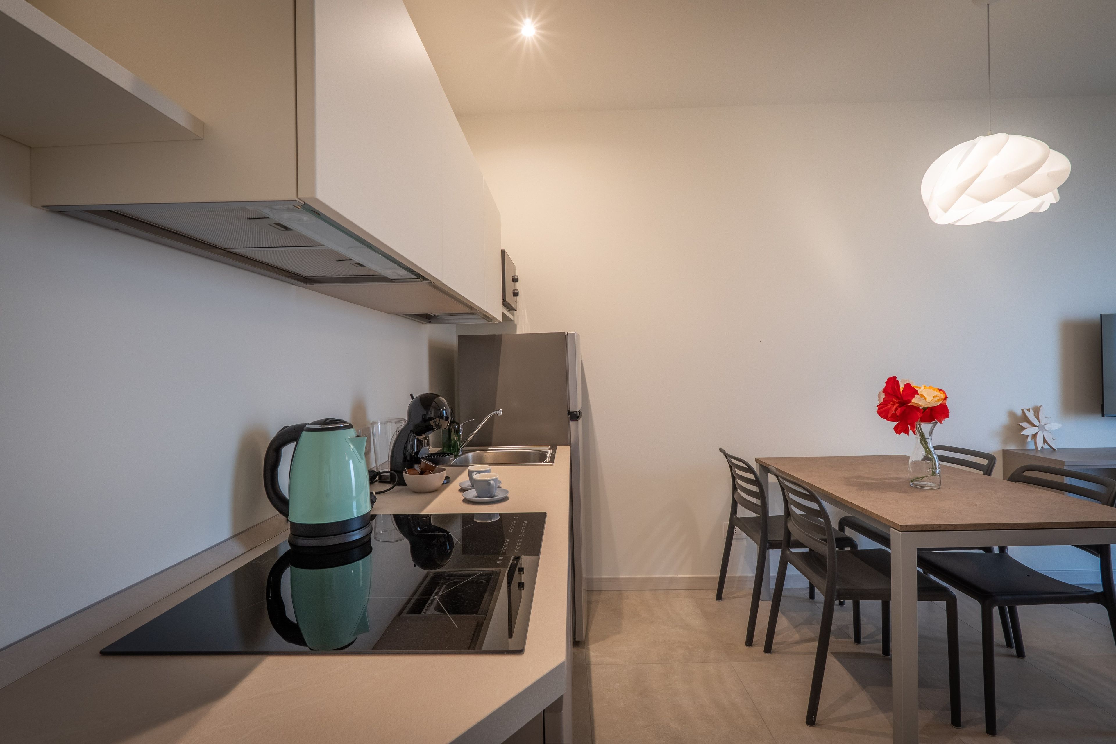 Une cuisine moderne équipée d’une cuisinière intégrée, d’une bouilloire et d’une machine à café. On y trouve une table à manger avec des chaises et un vase rempli de fleurs.
