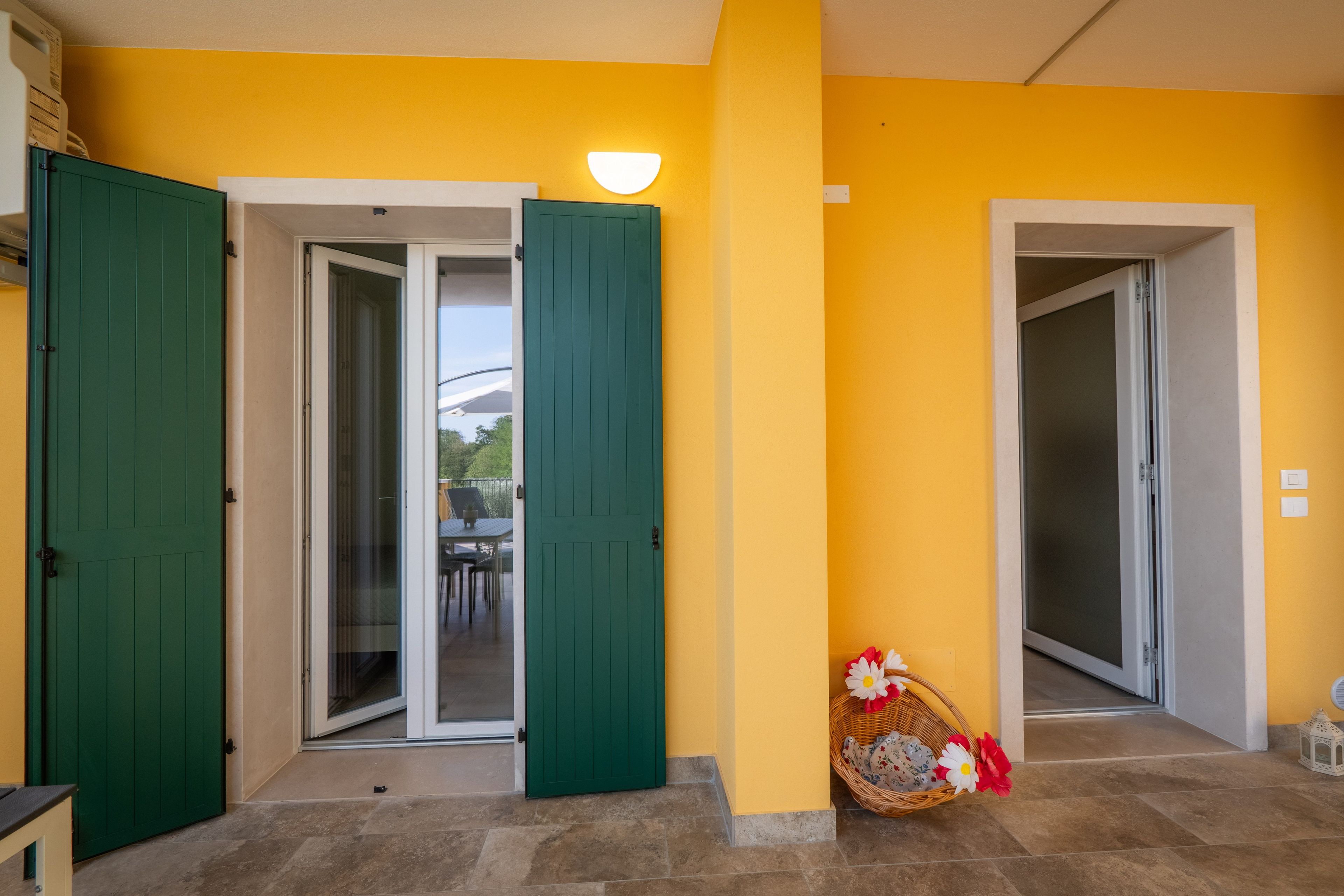 Un couloir avec deux portes vertes, dont l’une mène à une entrée avec une porte en verre et l’autre à une pièce. Un panier en osier rempli de fleurs est posé sur le sol.
