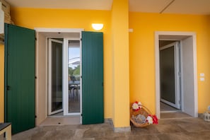 Un couloir avec deux portes vertes, dont l’une mène à une entrée avec une porte en verre et l’autre à une pièce. Un panier en osier rempli de fleurs est posé sur le sol.