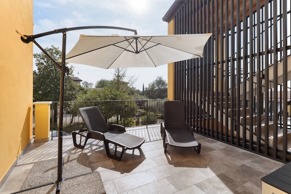 A balcony with two chairs and a large umbrella, a metal fence, and a building with vertical lines in the background.