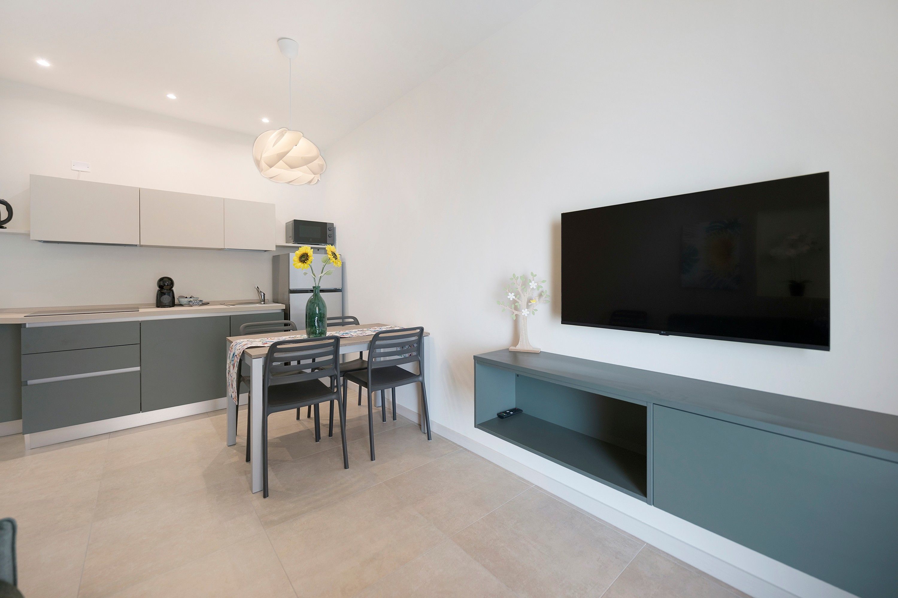 A modern kitchen with a dining area, a flat-screen TV mounted on the wall, and a ceiling light fixture.