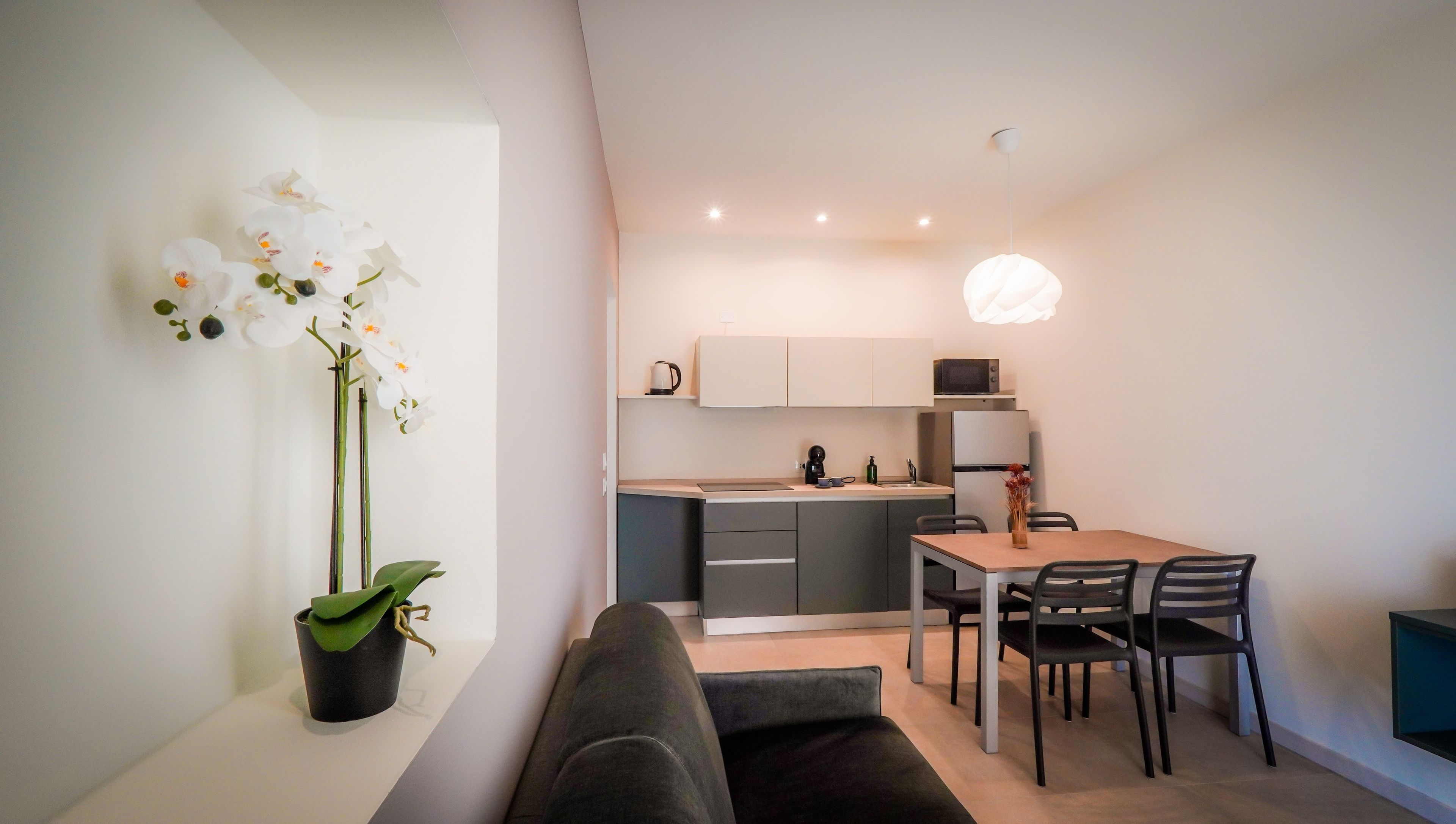 Une cuisine moderne avec des meubles blancs, une table à manger avec des chaises et une orchidée en pot sur la gauche.