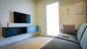 Un salon moderne avec un téléviseur à écran plat fixé au mur, un canapé gris avec une télécommande et une porte coulissante donnant sur un espace lumineux.