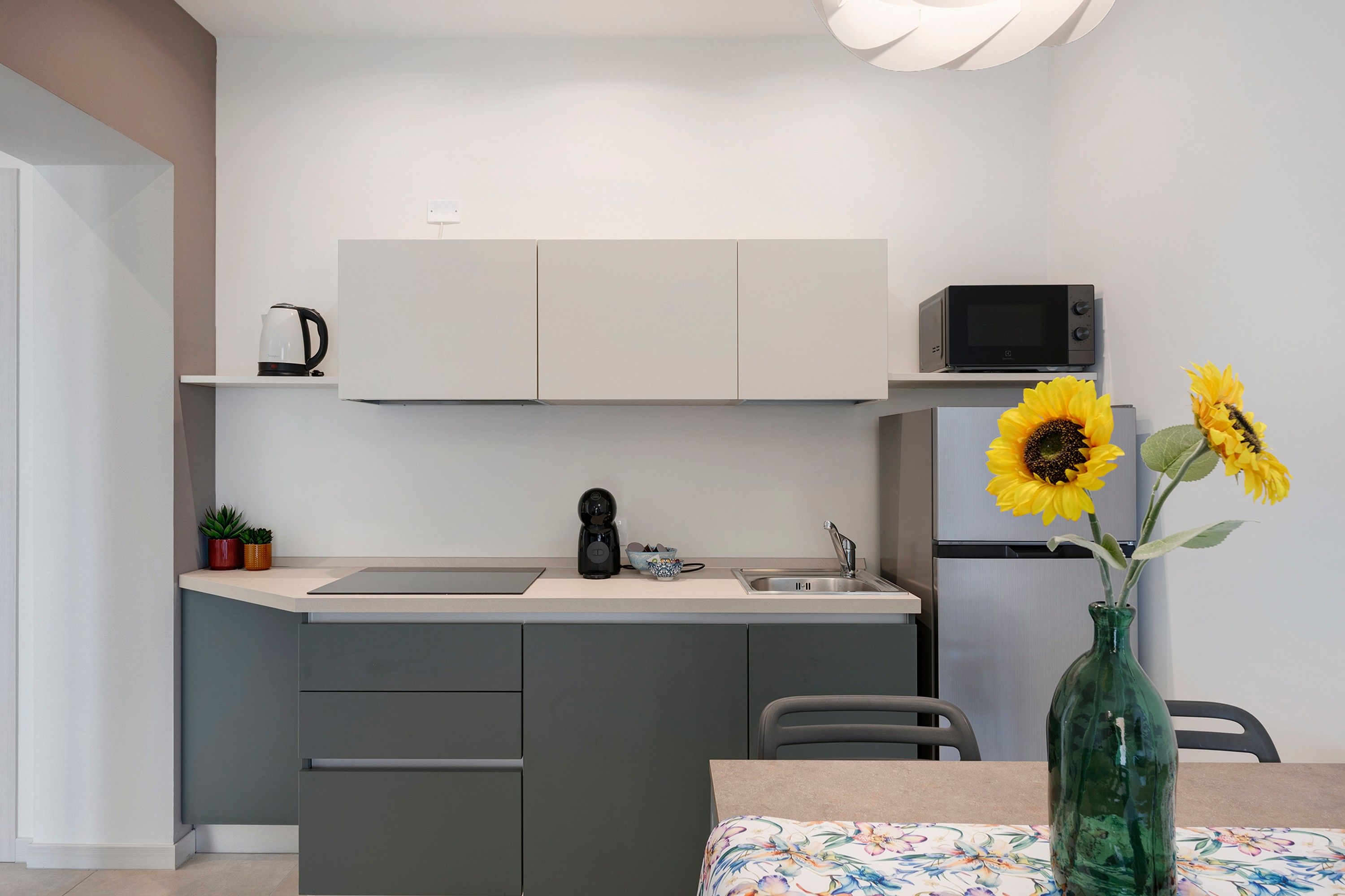 Una cocina compacta con microondas, hervidor y lavamanos. Hay una mesa pequeña con mantel floral y un jarrón con girasoles.