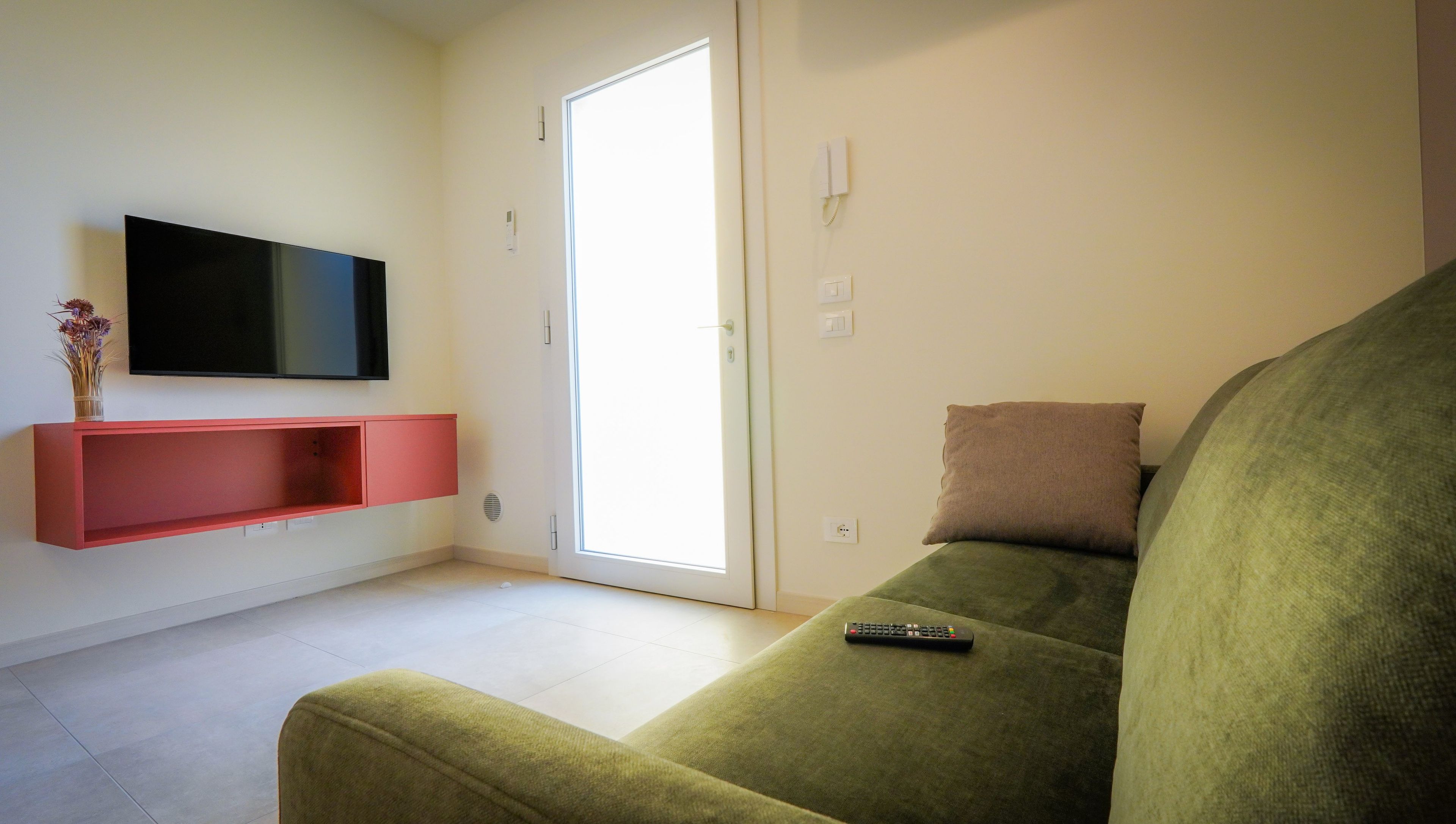 Un salon avec un canapé vert, un téléviseur à écran plat fixé sur une étagère rouge, une télécommande posée sur le canapé et une porte donnant sur un espace extérieur.