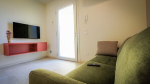 Un salon avec un canapé vert, un téléviseur à écran plat fixé sur une étagère rouge, une télécommande posée sur le canapé et une porte donnant sur un espace extérieur.