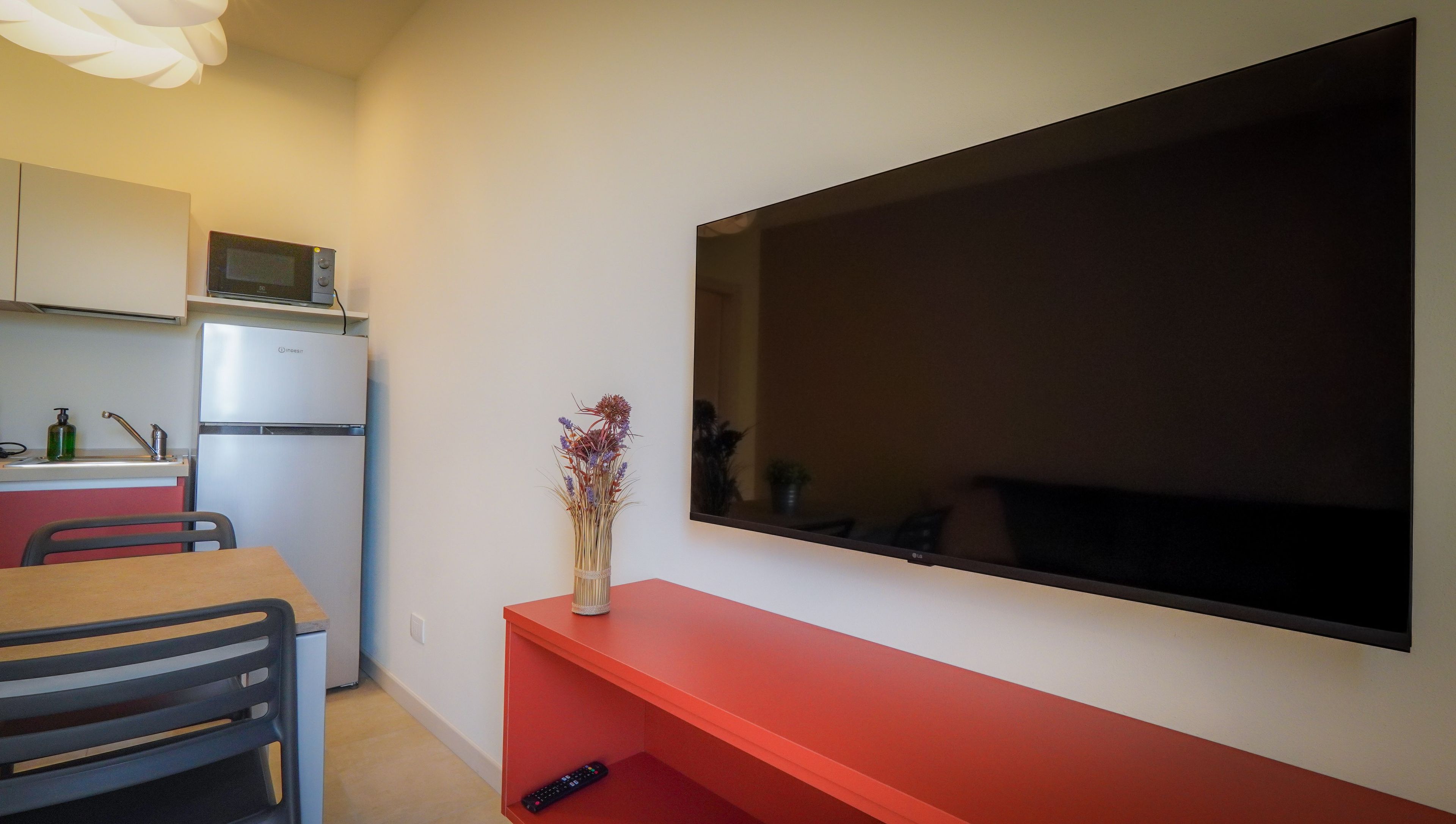 Una cocina compacta con refrigerador, microondas y un lavamanos pequeño. Un televisor de pantalla plana está montado en la pared sobre un estante rojo.