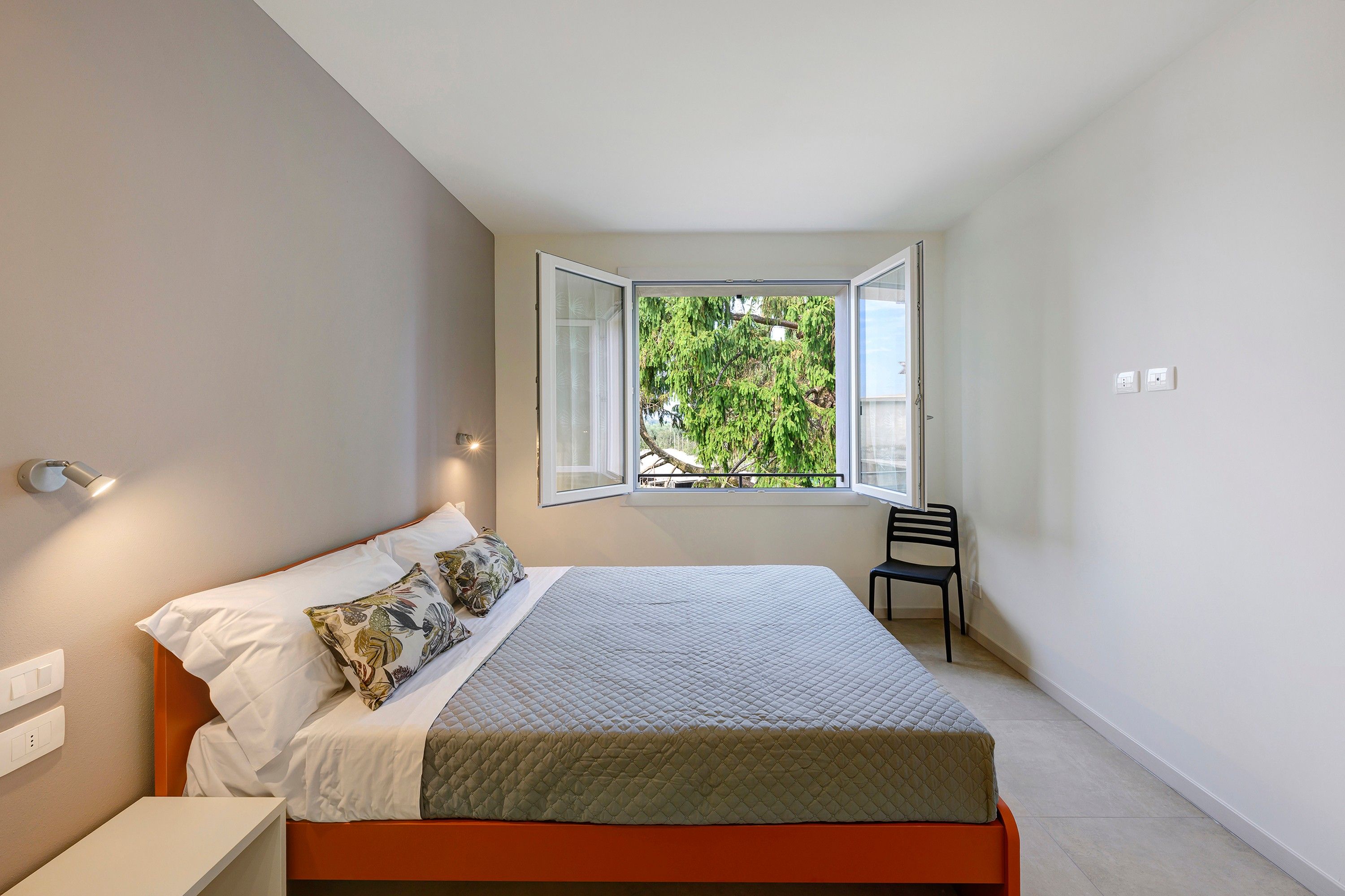 A bedroom with a bed, a chair, a window, and a wall-mounted light fixture.