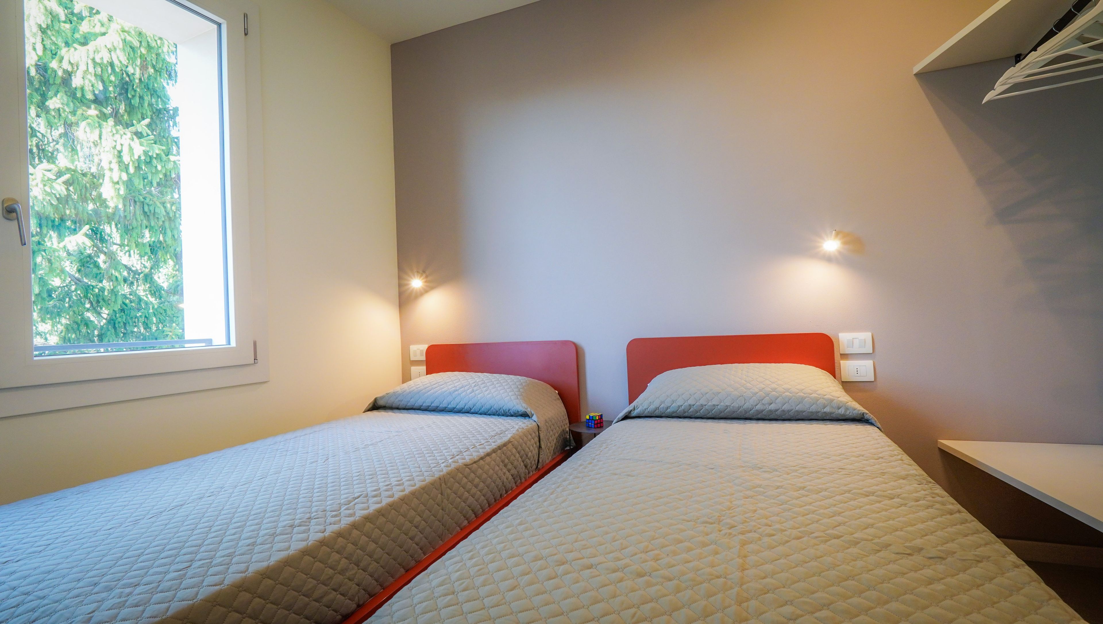 Une chambre avec deux lits, une fenêtre donnant sur la verdure et un luminaire fixé au mur.