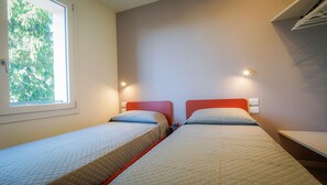 Une chambre avec deux lits, une fenêtre donnant sur la verdure et un luminaire fixé au mur.
