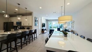 A modern kitchen with a dining area, featuring pendant lighting, a large dining table, and a view to the outdoors. - 6BR near Disney | Pool, spa & 2 game rooms (Kissimmee)
