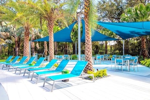 A poolside area with sun loungers and tables, surrounded by palm trees and shaded by blue canopies. - 8BR near Disney | Private pool, spa & game room (Kissimmee)
