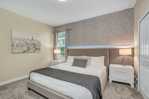 Un dormitorio con cama, mesitas de noche, lámpara, ventana con cortinas y un cuadro colgado en la pared.