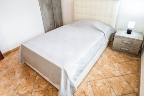 Una cama individual con una colcha gris en una habitación con suelo de baldosas y una mesita de noche con lámpara.