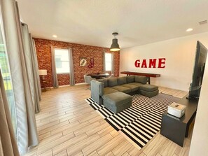 A modern living room with a sectional sofa, a black and white striped rug, a television, and a wall with the word 'GAME' in red letters. - 7BR near Disney | Private pool, spa & game room (Kissimmee)