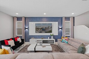 A modern living room with a flat-screen TV mounted on the wall, a black and red sofa, a beige sofa, and a white coffee table. - 11BR near Disney | Private pool, spa & cinema (Kissimmee)