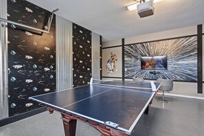 Ein Tischtennistisch in einem Raum mit Wänden zum Thema Weltraum, einem großen Fernsehbildschirm und einem an der Decke befestigten Projektor.