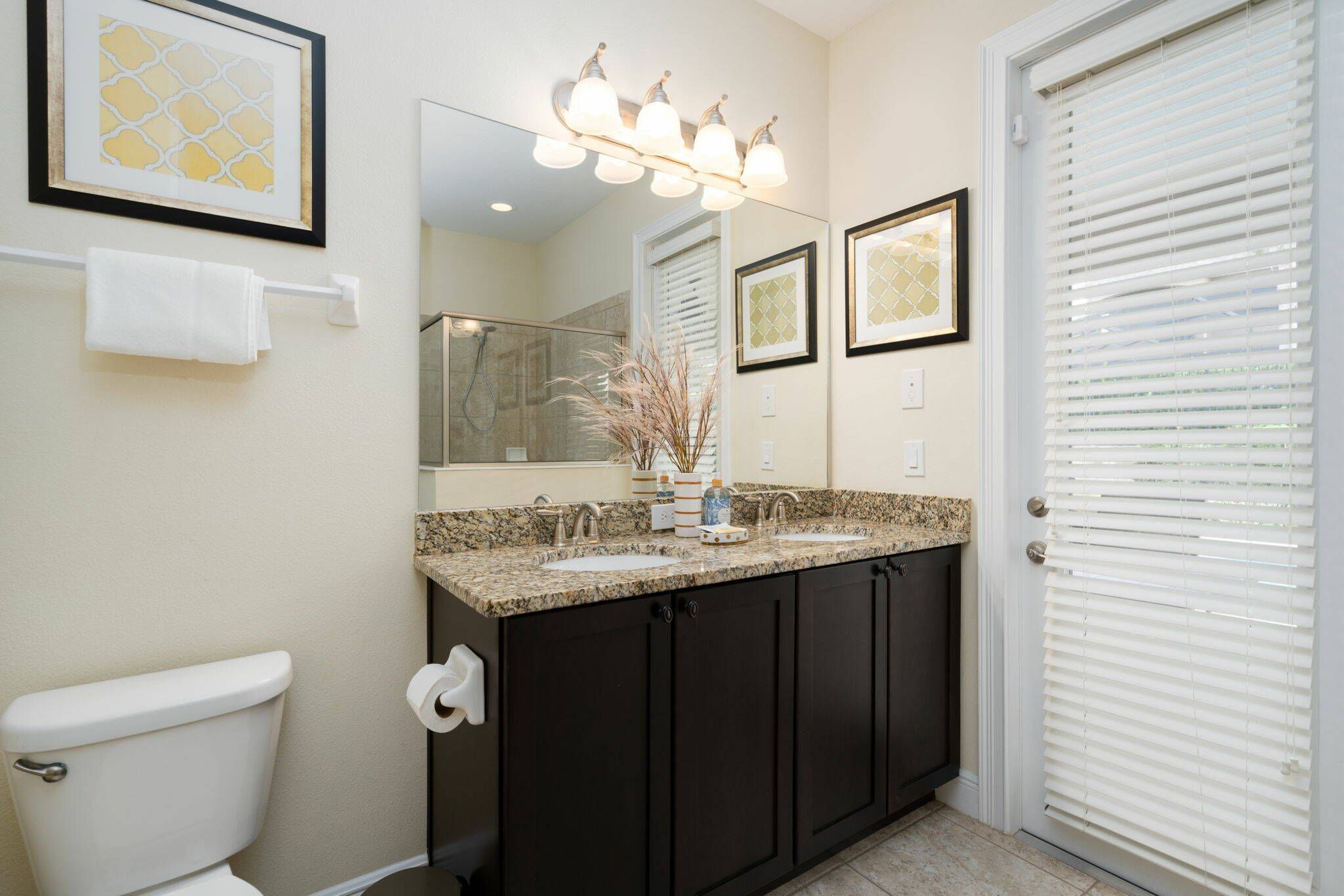 A bathroom with a toilet, dark cabinets, a granite countertop, a shower, and a window with blinds.