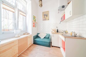 A modern kitchen with white cabinets, a teal sofa, and a window with blinds. - Kraków City Center Studio — Mezzanine (Kraków)