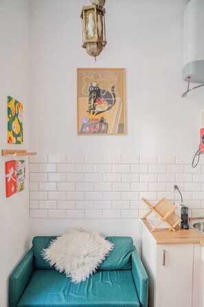 A compact living space with a teal sofa, a white fluffy pillow, a wall-mounted lamp, a framed poster, and a small kitchen area with a sink and a kettle. - Kraków City Center Studio — Mezzanine (Kraków)