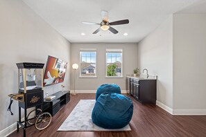 A room with a ceiling fan, a bean bag chair, a television on a stand, and a kitchen area with a sink. - 10BR near Disney | Private pool, spa & game room (Kissimmee)