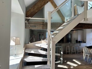 Eine Treppe mit Glasgeländern führt zu einer Erhöhung, ein Küchenbereich mit Barhocker und eine Lampe auf einem Tisch sind ebenfalls zu sehen.