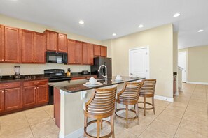 A modern kitchen with wooden cabinets, a black microwave, stainless steel appliances, and a bar area with wooden stools. - 5BR near Disney | Private pool, spa & game room (Kissimmee)