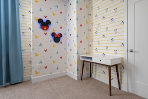 Quarto com porta branca, cortina azul, escrivaninha branca com gavetas e parede com estampas do Mickey Mouse.