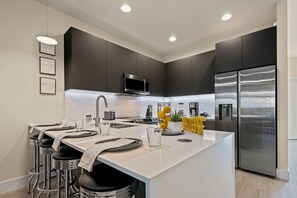 A modern kitchen with dark cabinetry, a stainless steel refrigerator, and a central island with bar stools. - 3BR townhouse near Disney | Pool & Mickey room (Clermont)