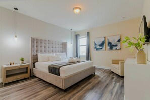 Un dormitorio moderno con cabecera capitonada, una mesita de noche con lámpara, cama con ropa de cama blanca, suelo de madera y dos cuadros abstractos en la pared.