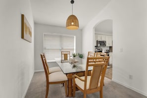 A dining area with a glass table, wooden chairs, a woven pendant light, and a view into a kitchen with white cabinets and appliances. - Terra Verde Oasis Private Pool (Kissimmee)