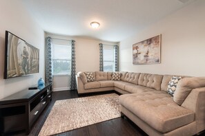 A living room with a sectional sofa, a flat-screen TV, a dark wooden entertainment unit, patterned curtains, and a large abstract painting. - 5BR near Disney | Pool, spa & 2 living rooms (Kissimmee)