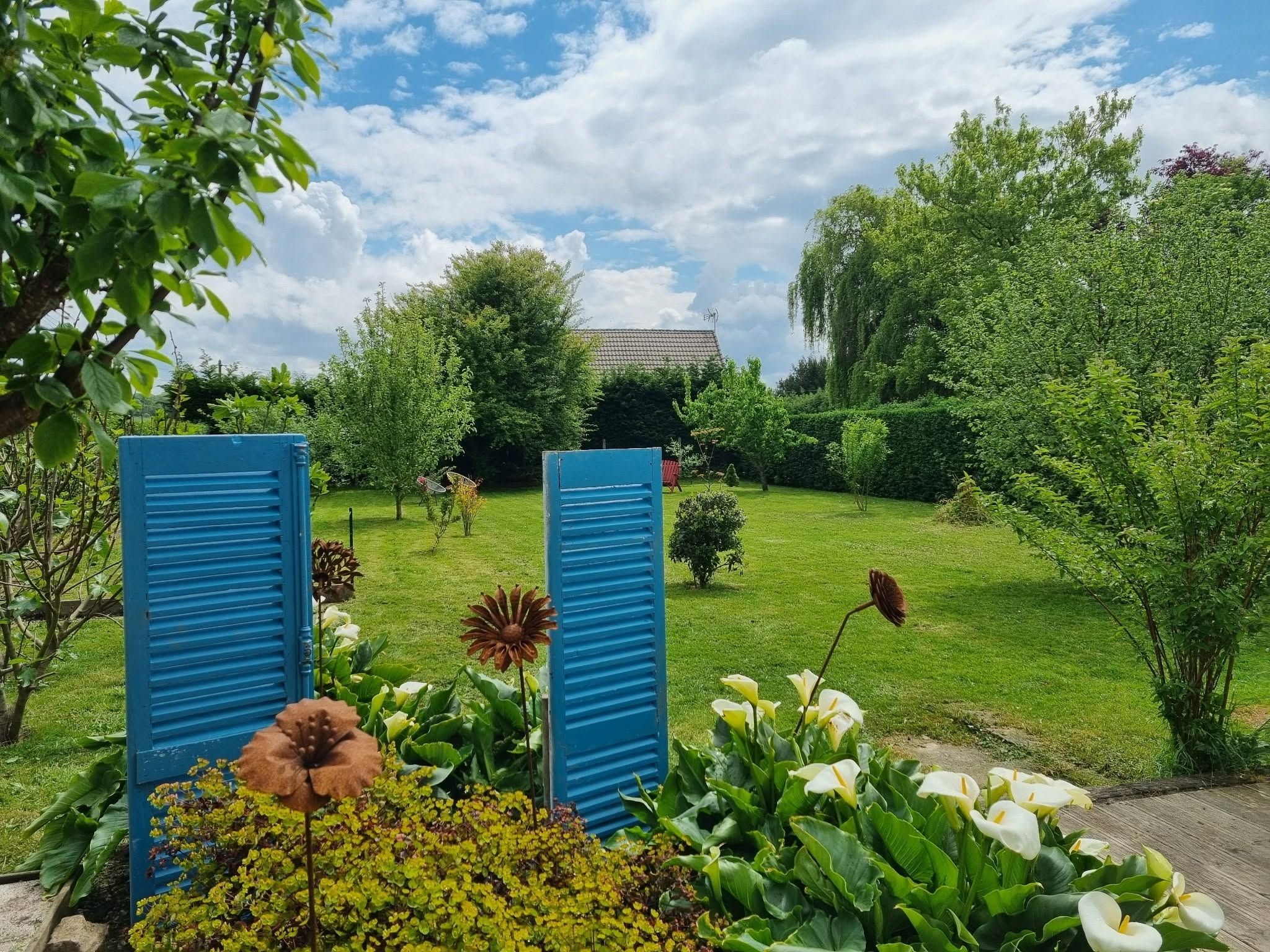 A garden with a lawn, a variety of plants, and a building with a tiled roof in the background.