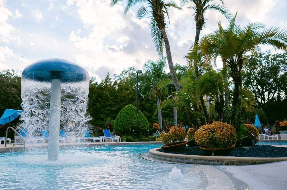 A swimming pool with a central water feature, surrounded by palm trees and lounge chairs.
