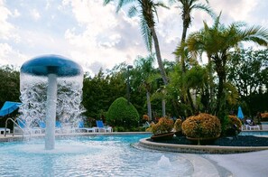 A swimming pool with a central water feature, surrounded by palm trees and lounge chairs. - 3BR townhome near Disney | Resort pool access (Kissimmee)