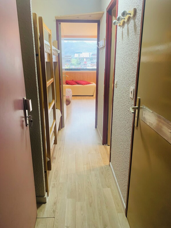A narrow hallway with wooden flooring leading to a bedroom with a window, a bed, and a red pillow. - Vrbo Property (Tignes)