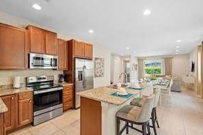 A modern kitchen with stainless steel appliances, wooden cabinets, and a dining area with a view. - 8BR near Disney | Pool, spa & game room (Kissimmee)