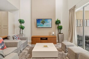 A modern living room with a flat-screen TV, two potted plants, a wooden TV stand, and a beige sofa. - 4 suites near Disney | Balcony & covered patio (Kissimmee)