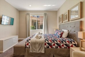 A bedroom with a bed, a bedside table, a TV mounted on the wall, and a window with curtains. - 4 suites near Disney | Balcony & covered patio (Kissimmee)