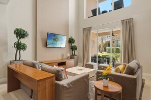 A modern living room with a sofa, coffee table, and a TV mounted on the wall. There are two potted plants and a view of the outdoors through large windows. - 4 suites near Disney | Balcony & covered patio (Kissimmee)