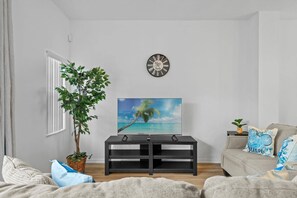 Ein Wohnzimmer mit großem Fenster, einem Blumentopf, einer Wanduhr, einem Flachbildfernseher und einer Couch mit Zierkissen.