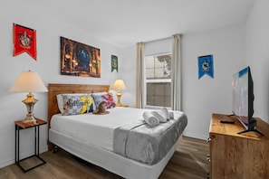 A hotel room with a bed, two bedside lamps, a television, and various wall decorations including flags and a painting. - 3BR near Disney | Mickey & Harry Potter rooms (Kissimmee)