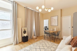 Een moderne woonkamer met een bank, een eethoek met een tafel en stoelen, een keuken op de achtergrond en een groot raam met gordijnen.