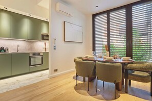 Eine moderne Küche mit grünen Schränken, ein Essbereich mit einem runden Tisch und Stühlen sowie große Fenster mit Jalousien.